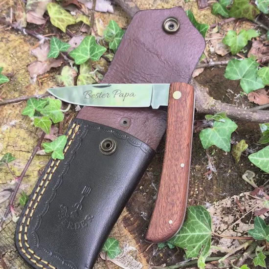 Hippekniep Taschenmesser mit Holzgriff und individueller Gravur liegt auf moosigem Holz in natürlicher Outdoor-Umgebung.